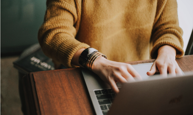 A person wearing a yellow sweater and multiple bracelets is typing on a laptop. The image is taken from an angle above the person's left shoulder, showing the hands on the keyboard and part of the laptop screen. The background is blurred, with a hint of a book and possibly a desk or table.